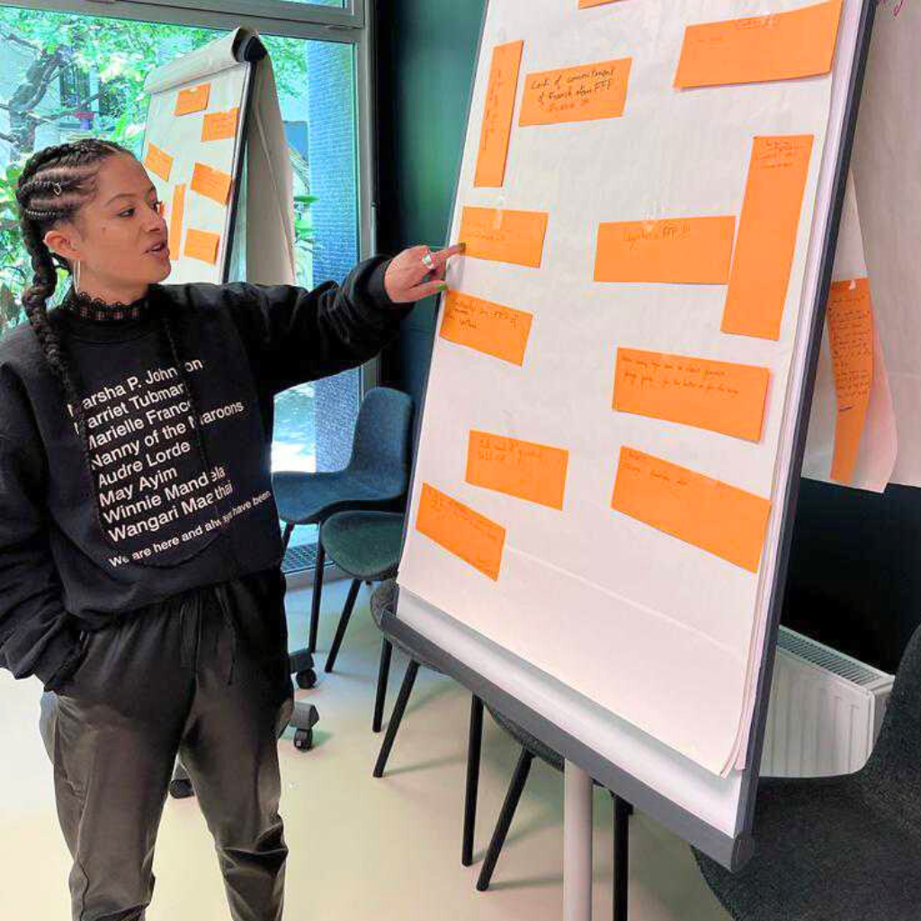 Project Manager Sheena Anderson, leading a workshop on Feminist Foreign Policy in Paris in 2023, for Amnesty International. She is standing in front of a white board with orange stickers. She is pointing at one of the stickers while speaking.